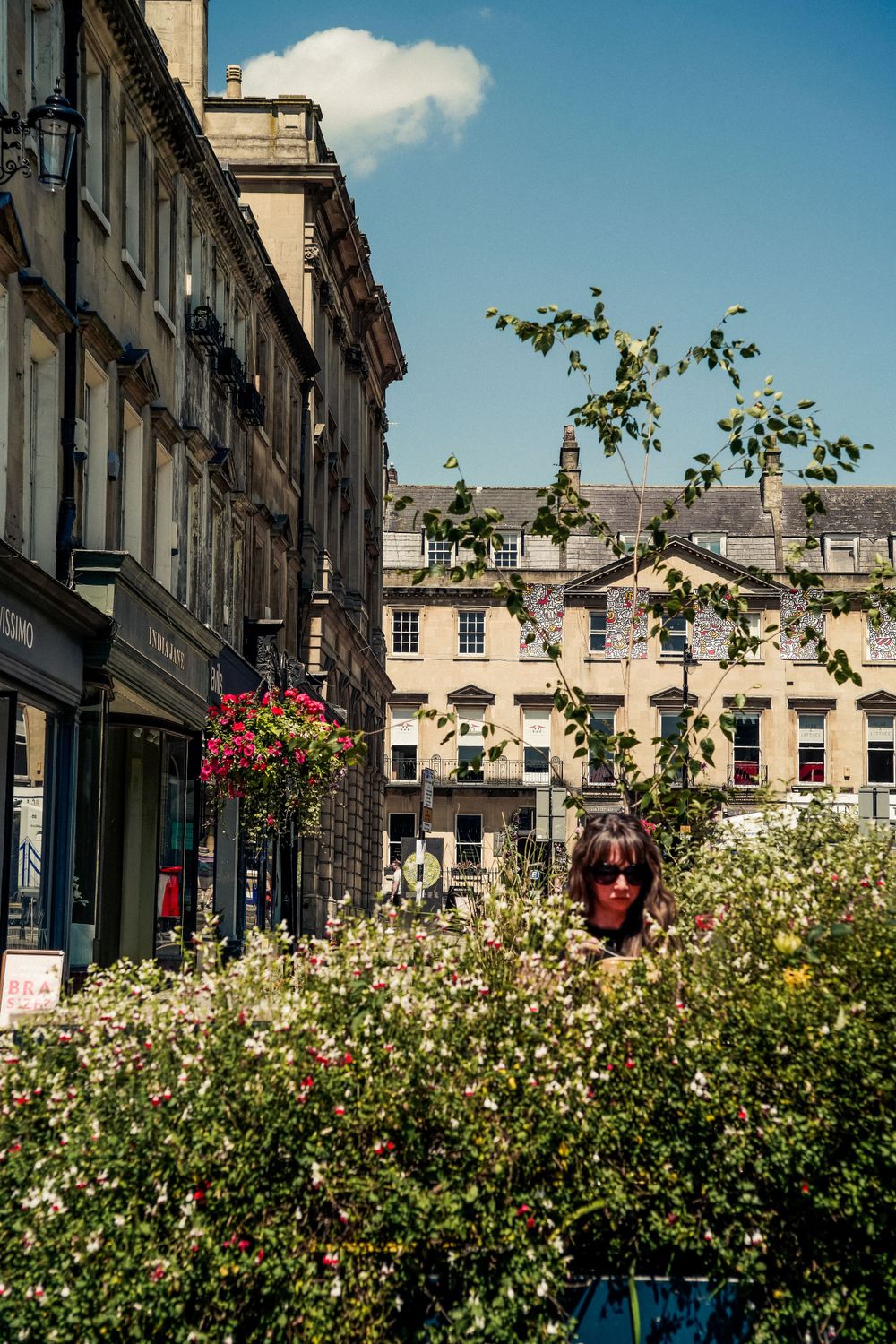 Historic stone buildings with flowering bushes and hanging baskets on a sunny day in Bath.