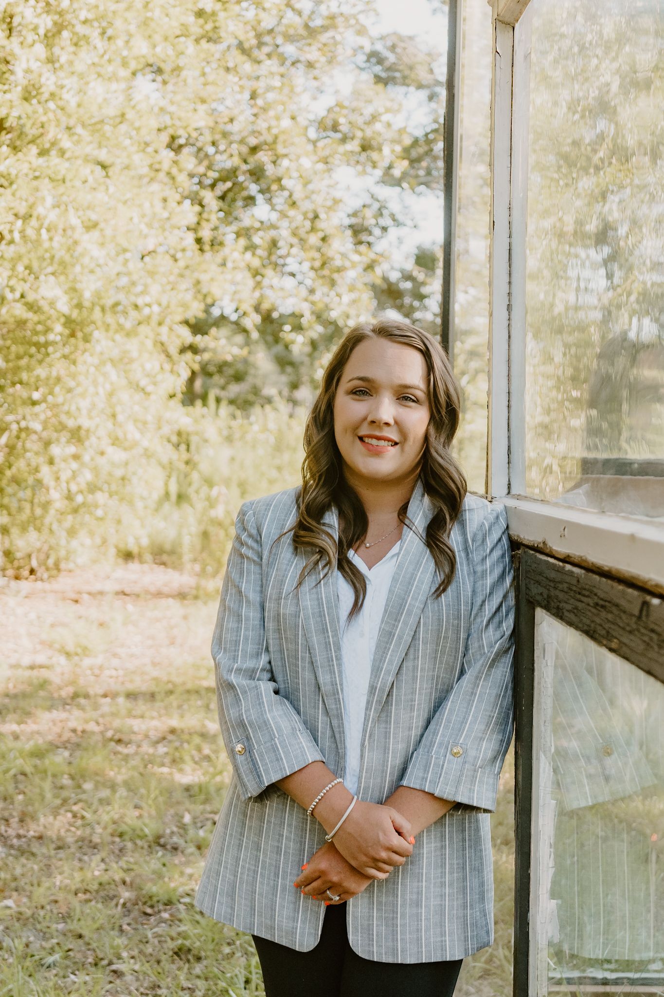 A person in a gray pinstriped blazer and white shirt stands outdoors against a greenhouse wall on a sunny day.