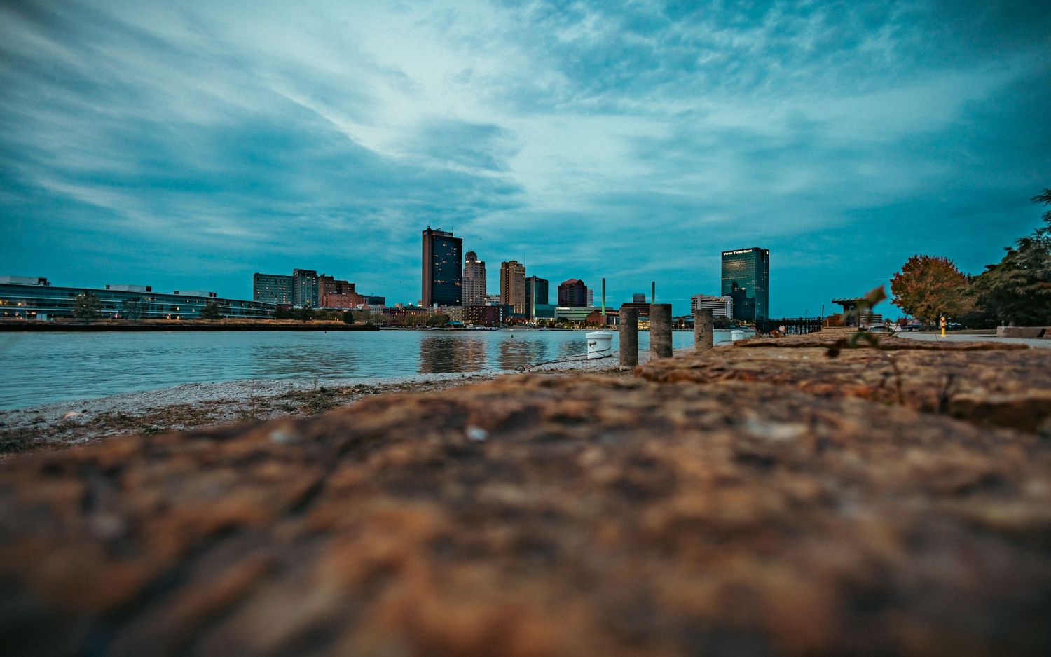 Urban skyline reflected in water with rocky foreground under dramatic blue sky.