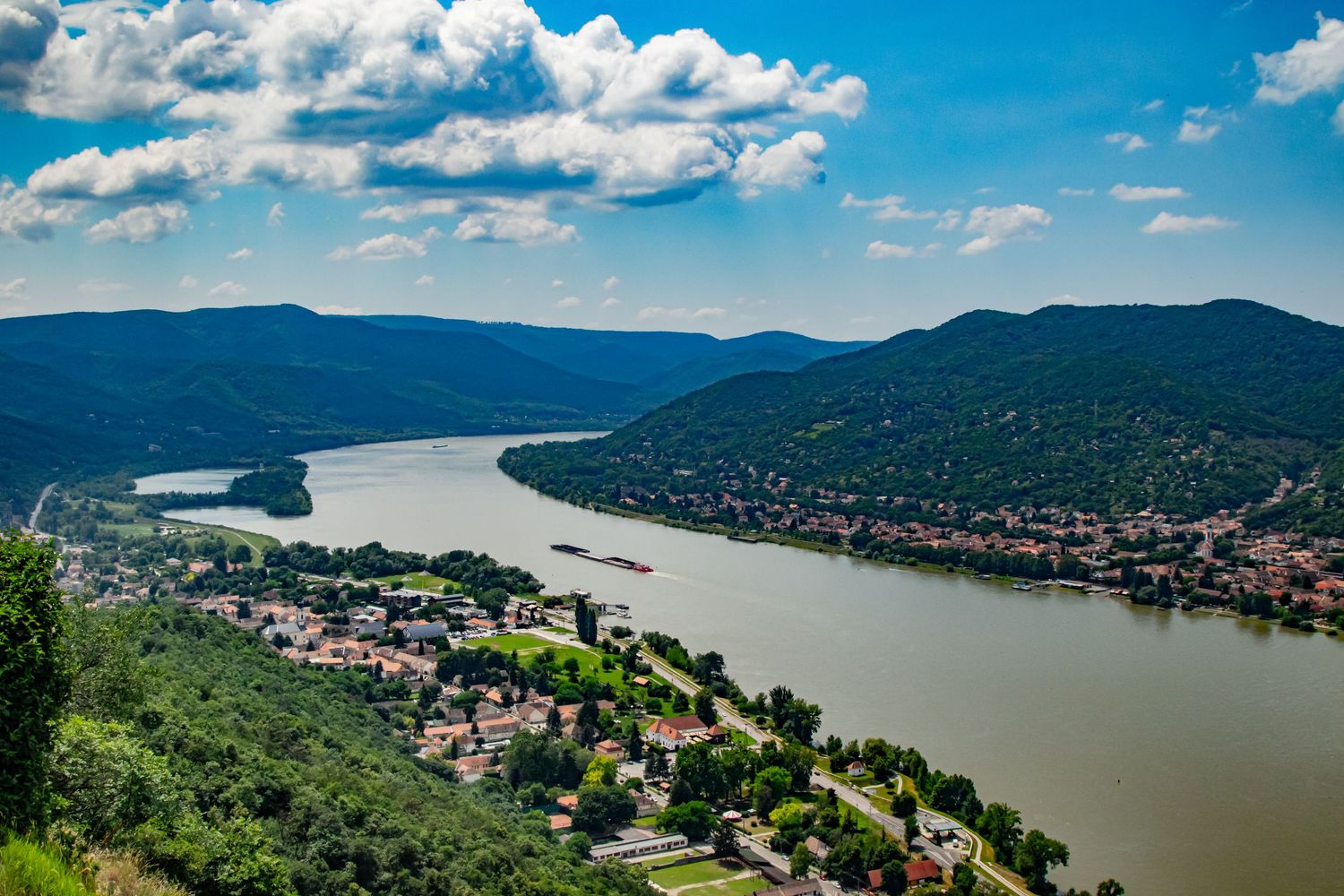 Panoramic view of a winding river valley with residential buildings nestled between forested hills.