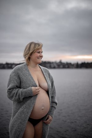 Gravidfotografering vid vatten. Kvinna står med grå kofta och magen synlig, vatten i bakgrunden