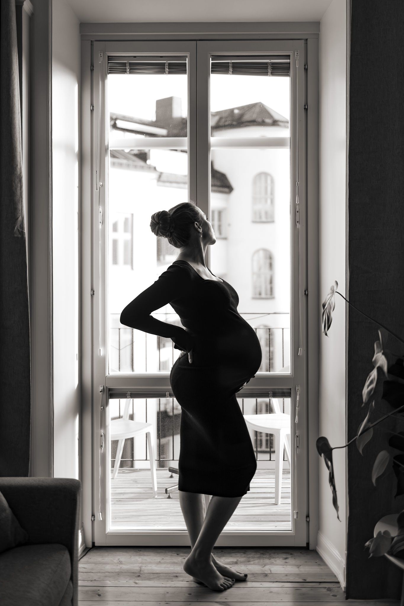 Svartvit bild i silhuette på gravid kvinna. Står i fönstret med svart klänning och tittar ut.