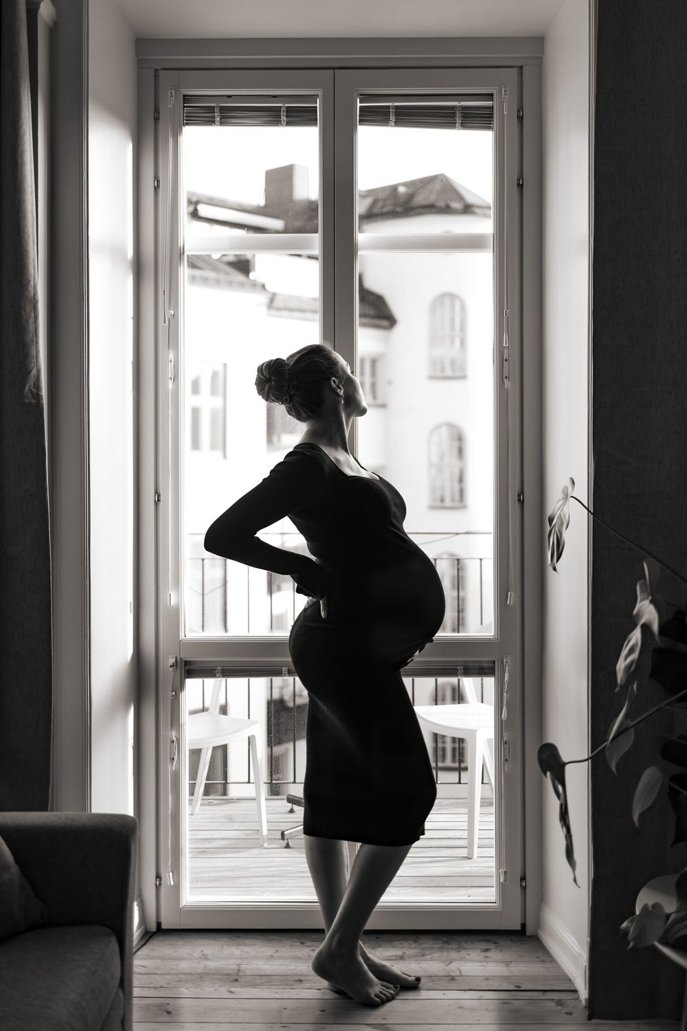 Svartvit bild i silhuette på gravid kvinna. Står i fönstret med svart klänning och tittar ut.