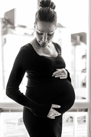 Svartvit bild på gravid kvinna i motljus. Hon tittar ner, håller händerna på magen i en svart klänning.