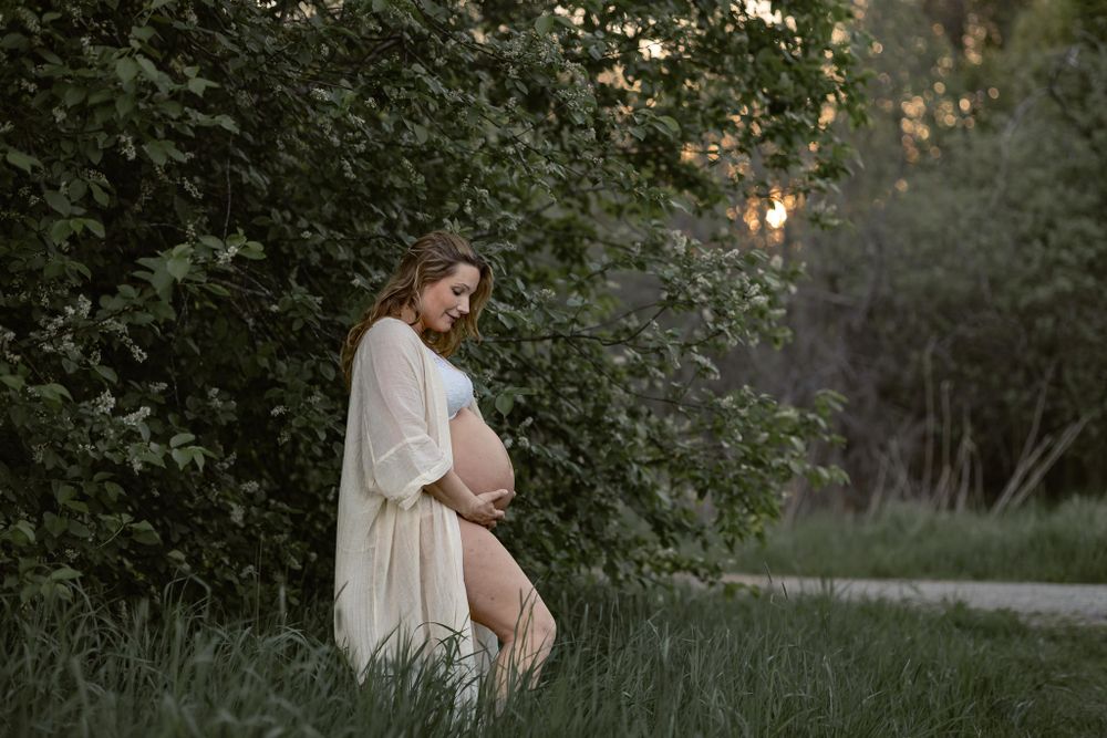 Gravidfotografering utomhus i solnedgång. Kvinna står vid träd i vit kimono, ser ner på magen. Solen skiner igenom bladverket.