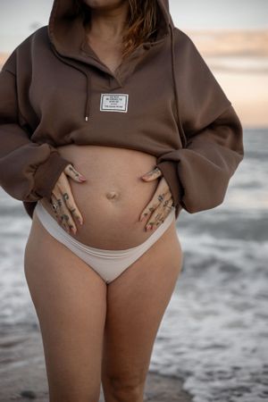 Gravidfotografering på stranden. Närbild på mage där kvinnan har en brun luftröja och håller händerna vid sidan av magen. Vågor i bakgrunden