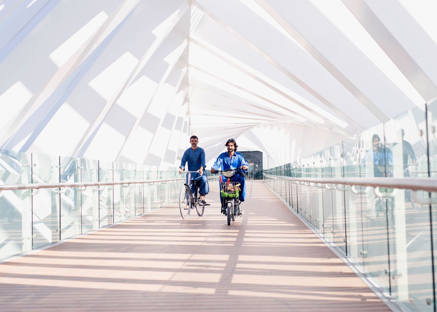 Two cyclists ride their bikes on a modern covered bridge walkway with geometric white architecture and glass panels.