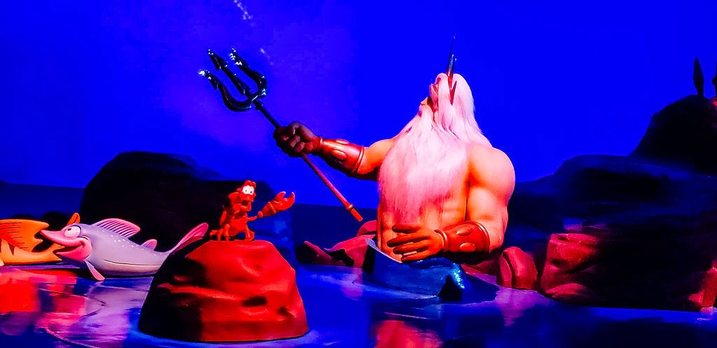 A reddish-pink figure emerges from dark water against a vibrant blue background creating a surreal theatrical scene.