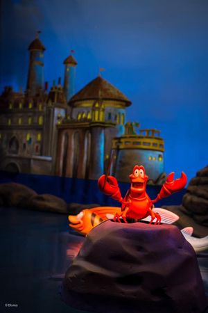 Sebastian the crab from The Little Mermaid performs in front of a castle backdrop at a Disney theme park attraction.