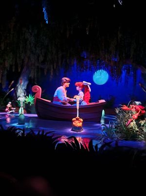 Two figures share a romantic moonlit boat ride surrounded by glowing blue lights and lush vegetation in a dark enchanted setting.