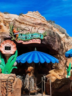 Entrance to a themed attraction "Little Mermaid" with a blue umbrella and rock formation facade against bright blue sky.