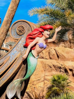 A little mermaid statue with red hair and green tail perches on rocky cliffs against a bright blue sky at a theme park.