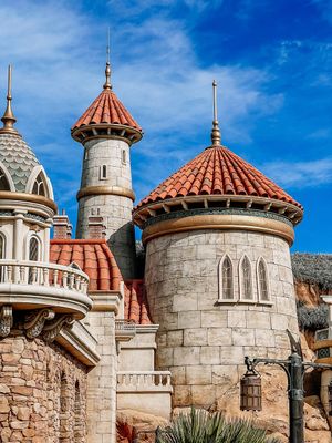 Architectural details of a fantasy castle with white stone towers and terra cotta domed roofs against a blue sky.