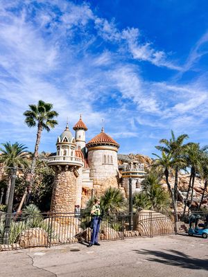 Panoramic view of a Mediterranean palace with domed towers, palm trees, and stone walls against a bright blue sky.