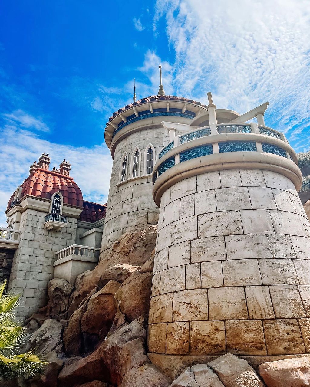 A white stone castle turret with circular tower and ornate architectural details against a bright blue sky with wispy clouds.