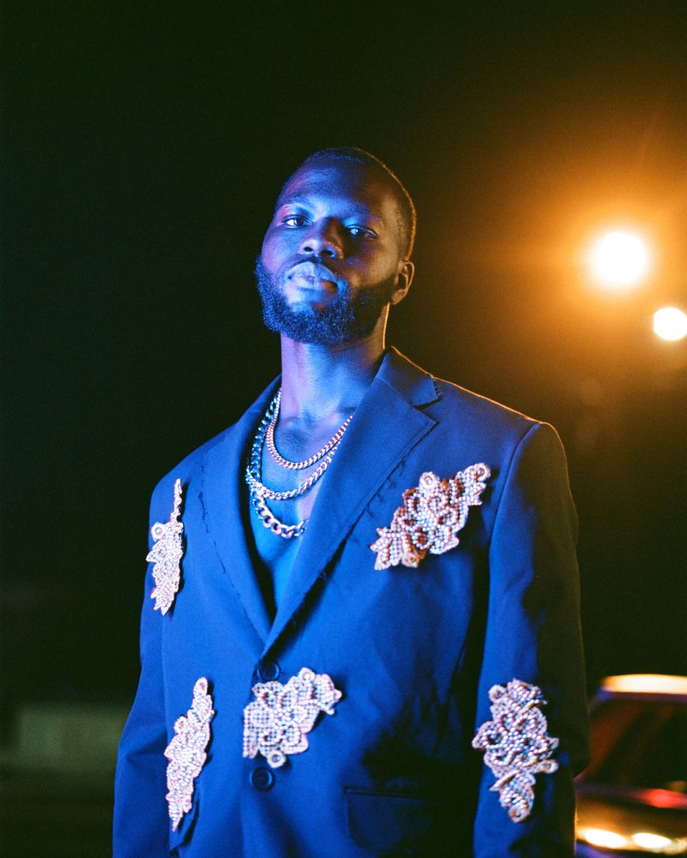 Person in blue embellished blazer illuminated by warm lighting during nighttime photoshoot.