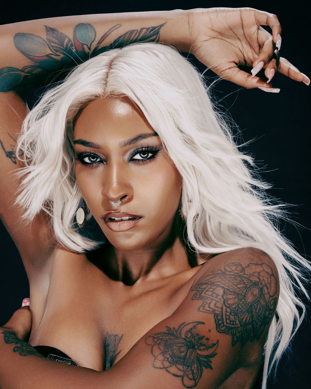 Editorial beauty shot featuring platinum white hair, dramatic makeup and visible floral tattoo artwork.