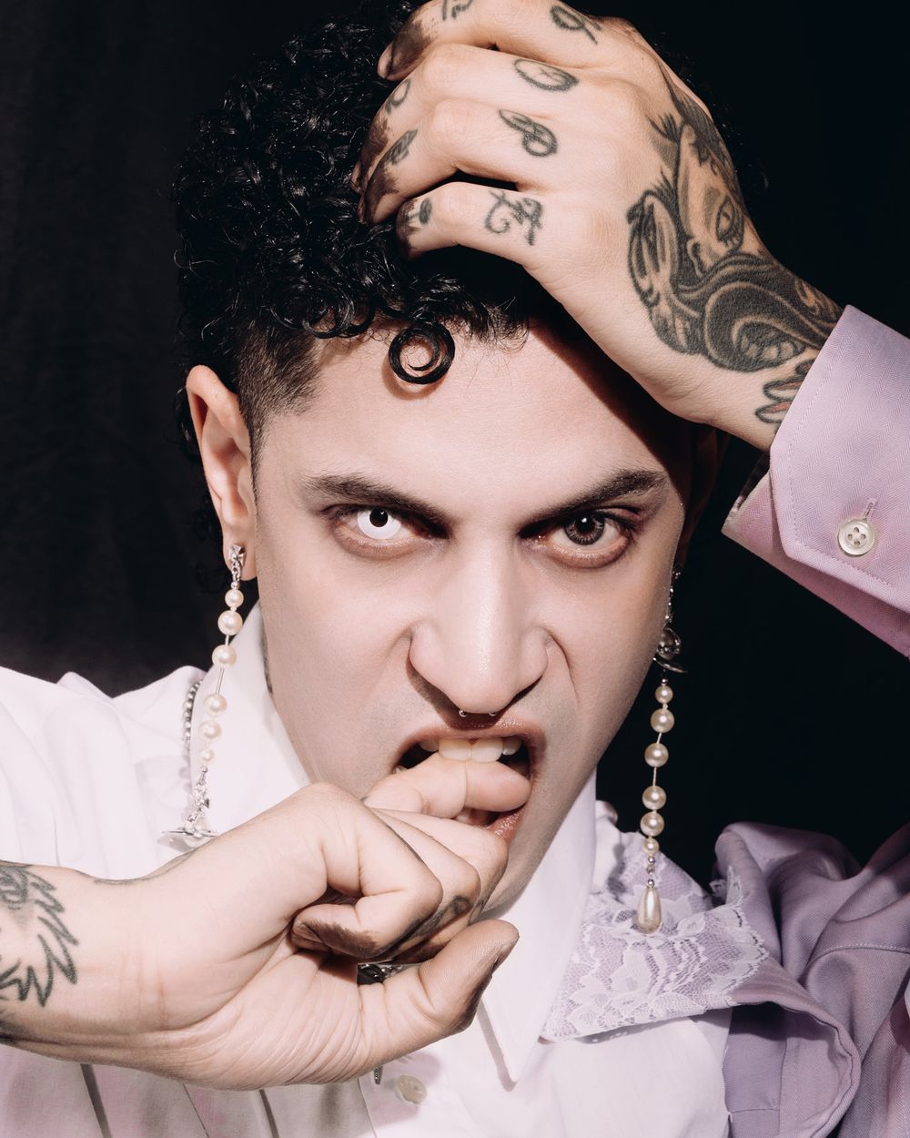 Portrait of someone posing in pink clothing with tattoos and pearl jewelry against a dark background.