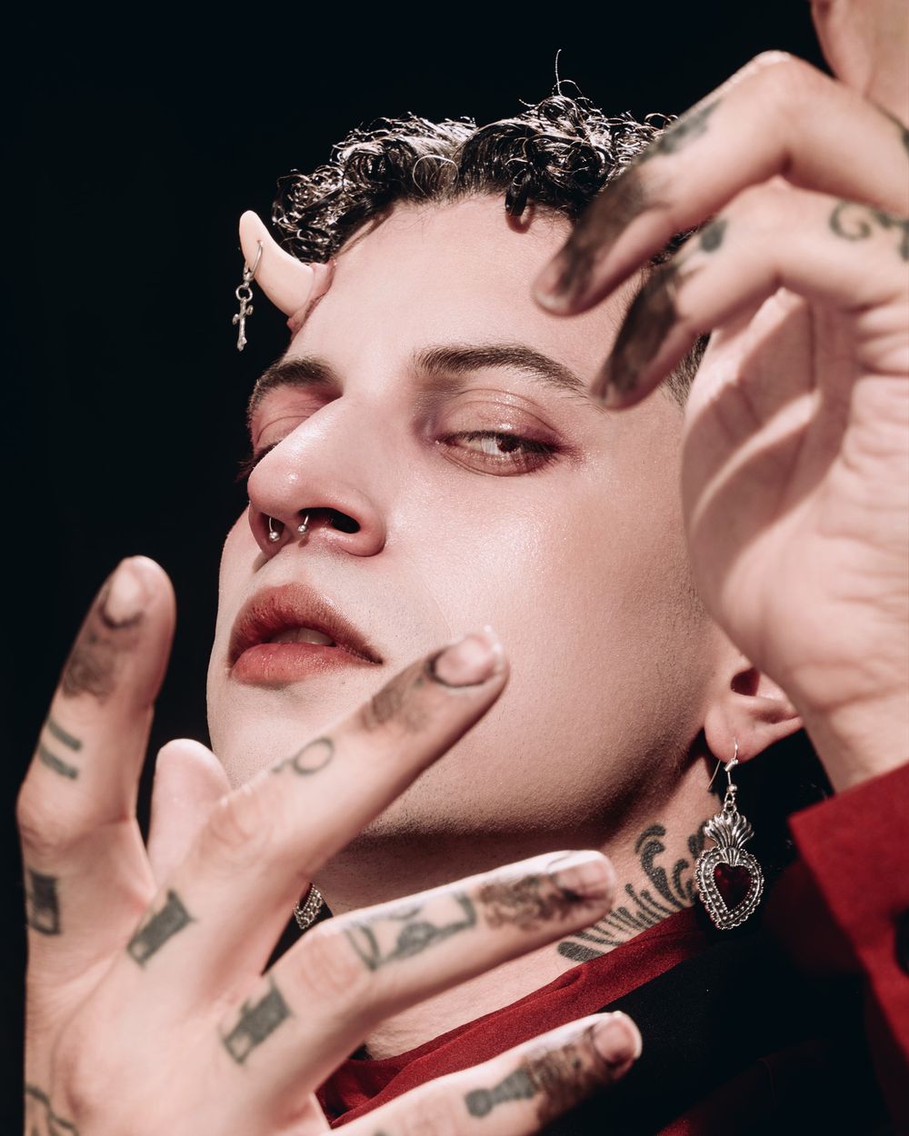 Close-up artistic portrait showing curly dark hair, red lipstick, tattooed fingers and silver earrings against a dark background.