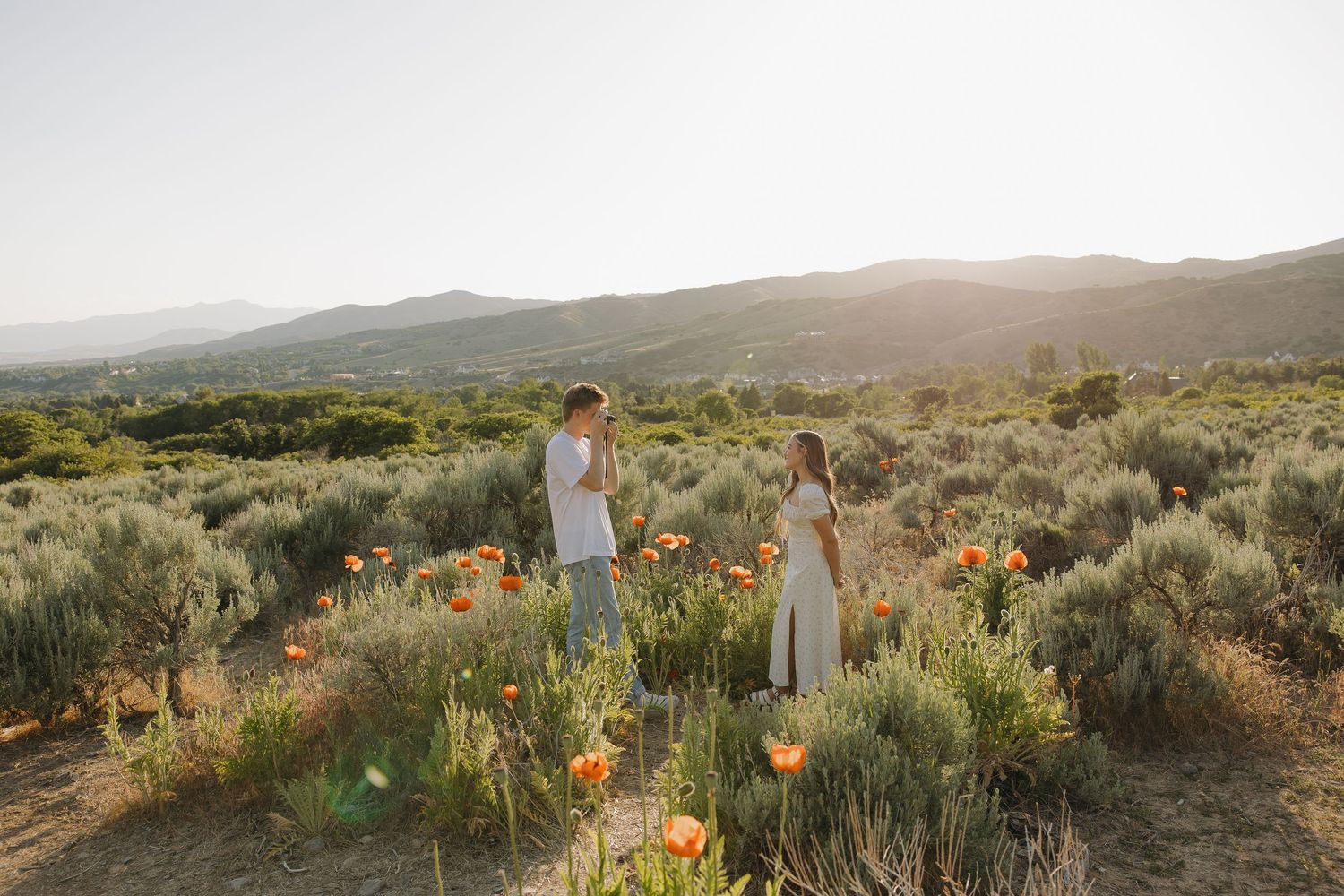 Couple standing in a sunlit field of orange wildflowers with rolling hills in the background.