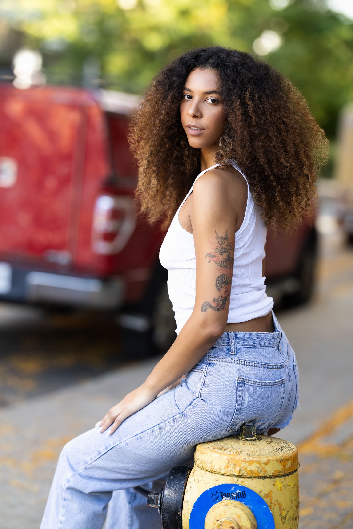 Woman in white tank top and light jeans sits on yellow fire hydrant outdoors with curly hair and tattoos.