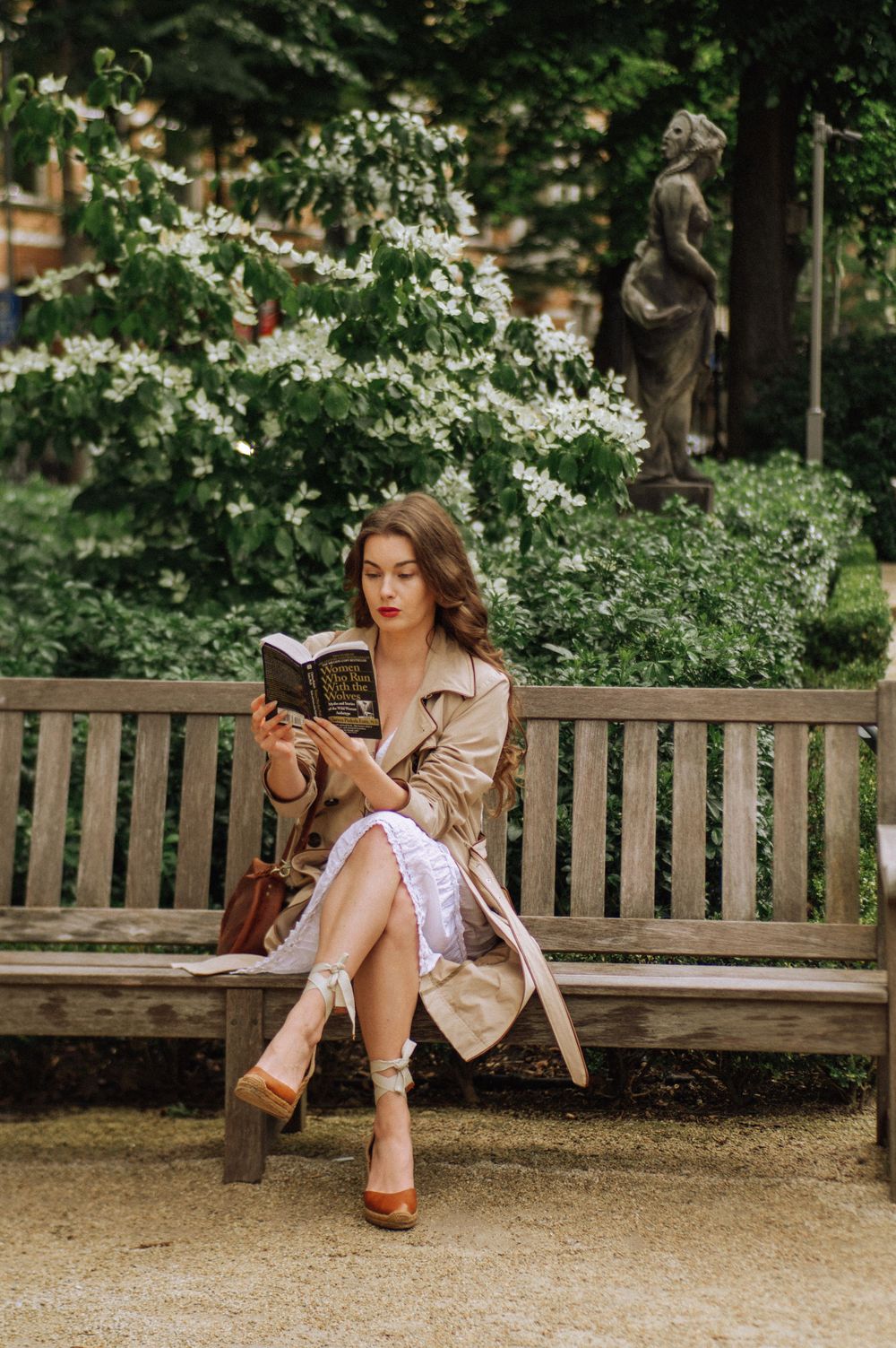 Woman in white dress and tan jacket sits casually on wooden park bench surrounded by greenery.