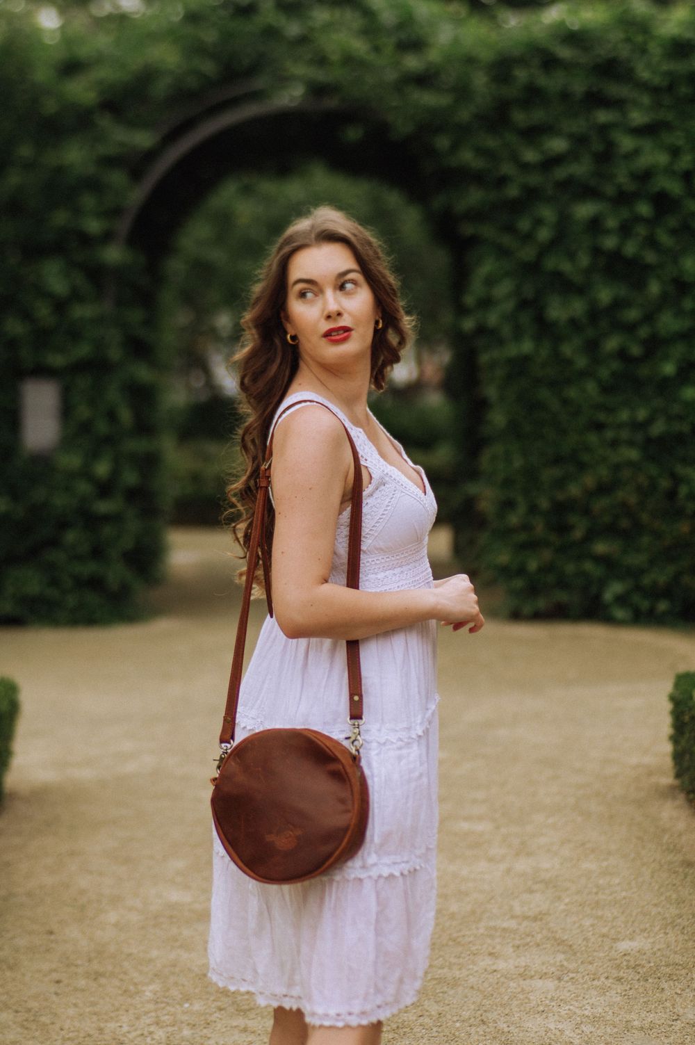 Woman in white eyelet dress carries brown leather crossbody bag through garden pathway.