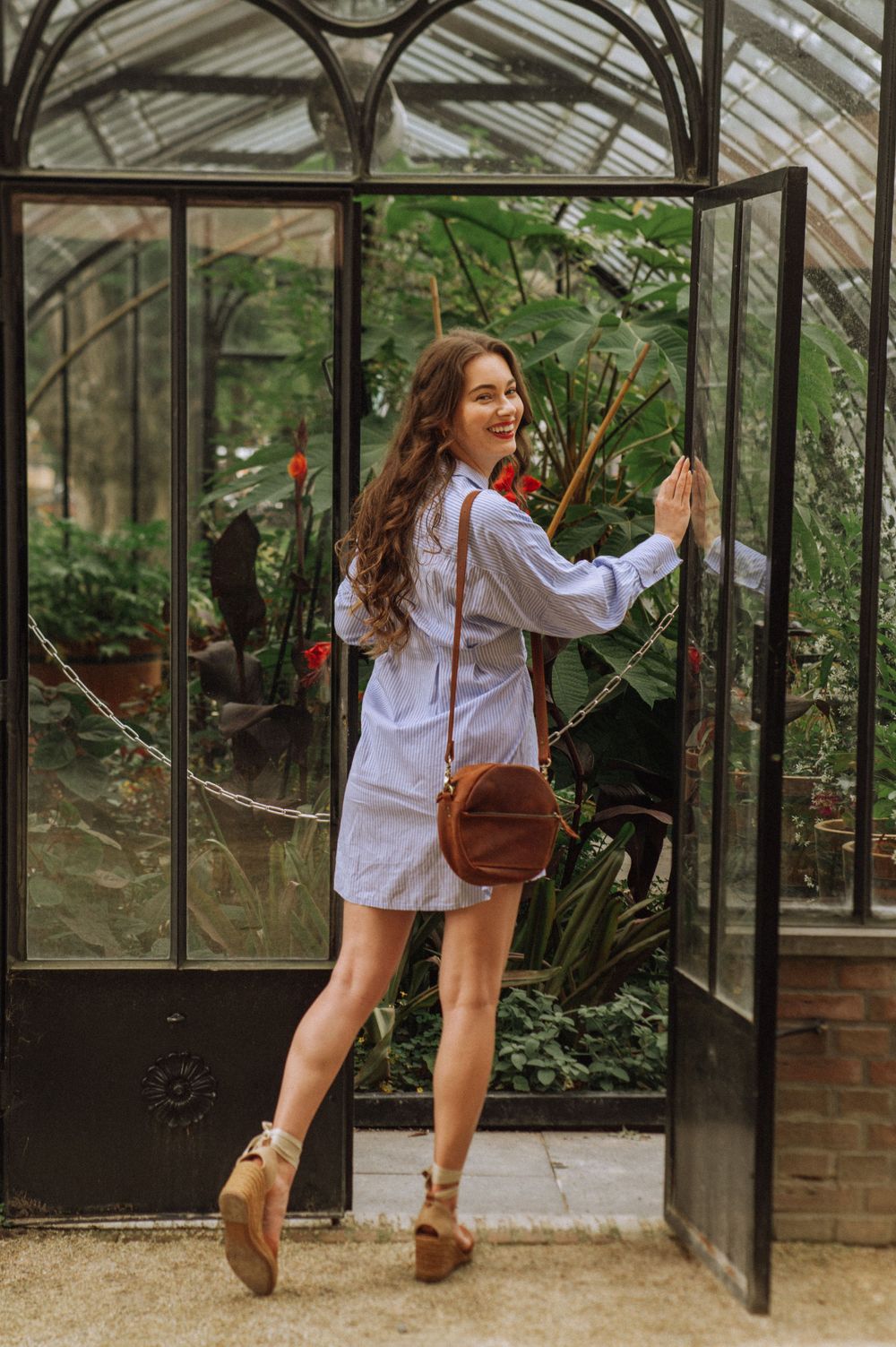 Woman in blue shirt poses playfully by glass greenhouse structure surrounded by plants.