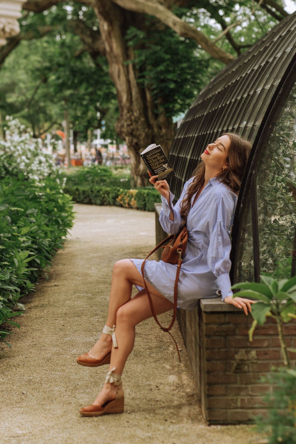 Woman relaxes on curved garden bench wearing blue shirt and brown shoes in lush park setting.