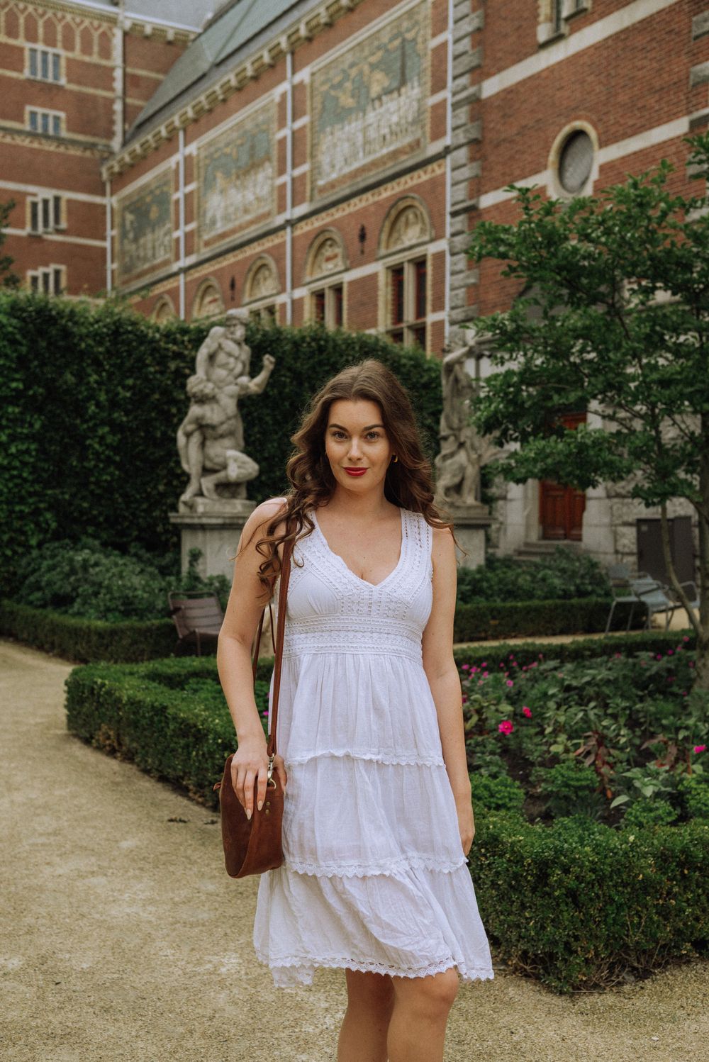 Woman in a white summer dress poses near a classic brick building with manicured gardens.