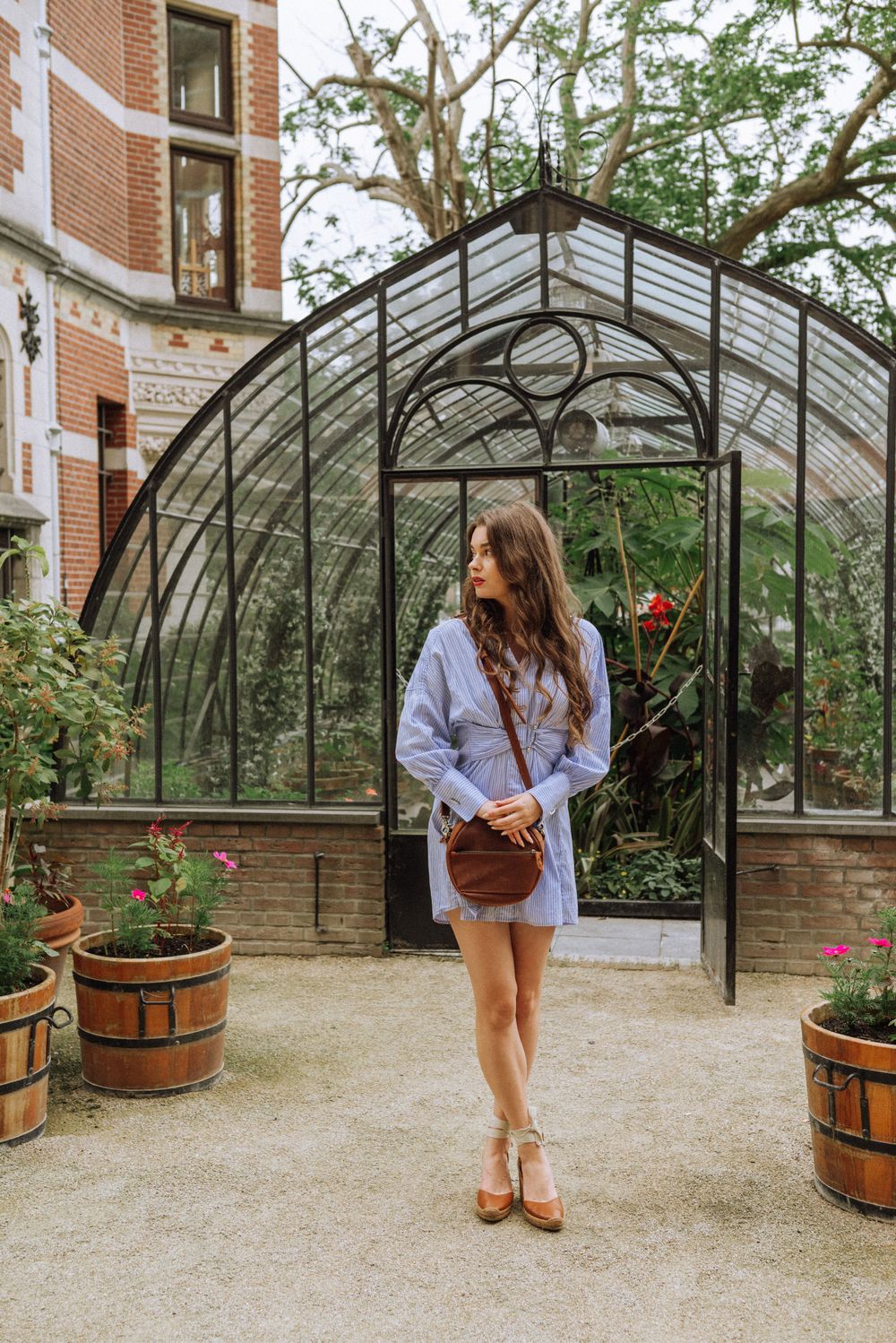 Woman poses playfully near greenhouse with wooden barrel planters and brick walls.