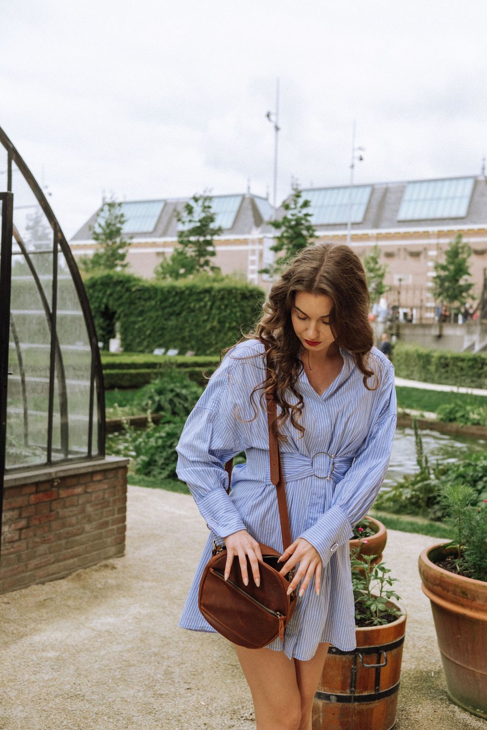 Woman in blue shirt examines plants near wooden barrel planters in greenhouse setting.