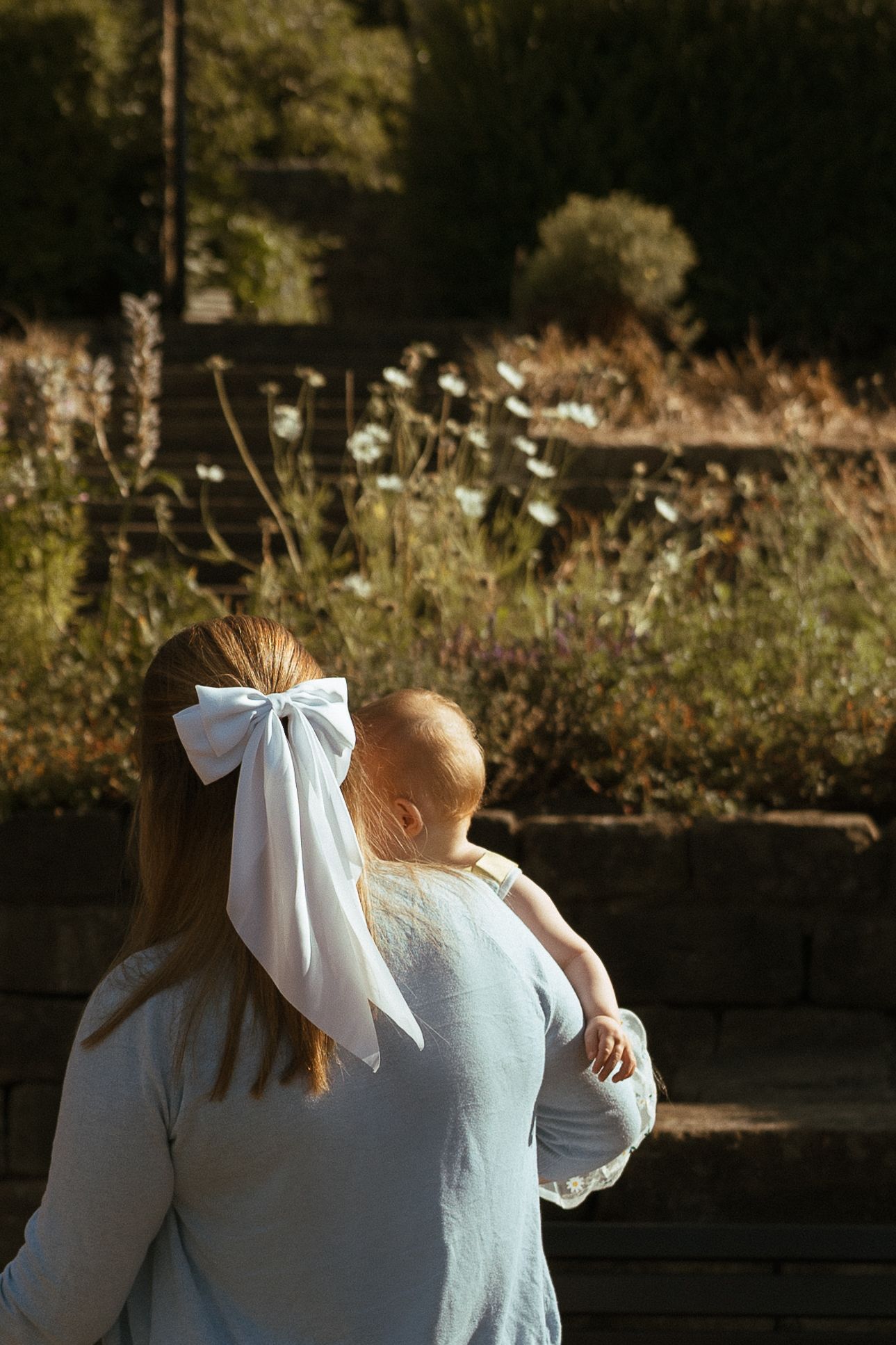 A woman with a white bow in their hair holds a baby while looking at wildflowers during a family photo session at Kirkstall Museum Gardens.