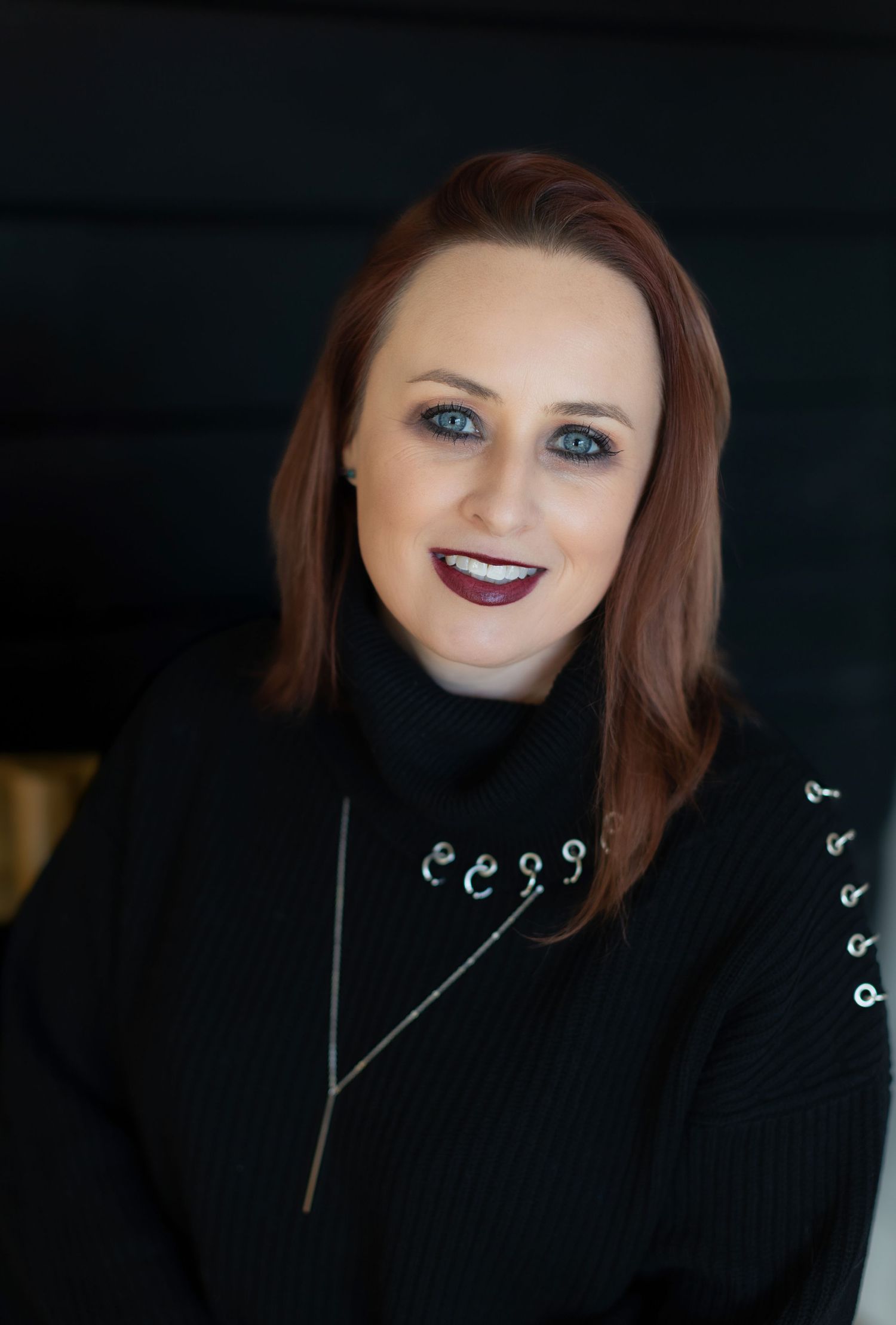 Professional portrait with dark background showing someone wearing a black turtleneck sweater and silver necklace.