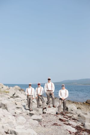 A group wearing white shirts and suspenders stands together on rocky coastal terrain overlooking the ocean.