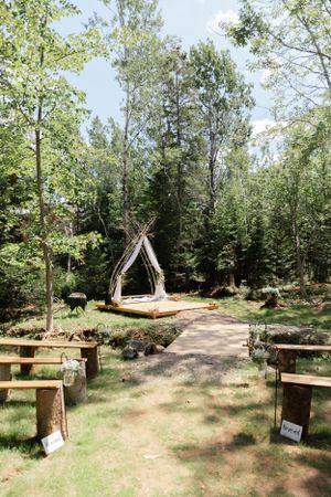 Rustic outdoor wedding ceremony setup with wooden benches and a triangle arch in a forest clearing.