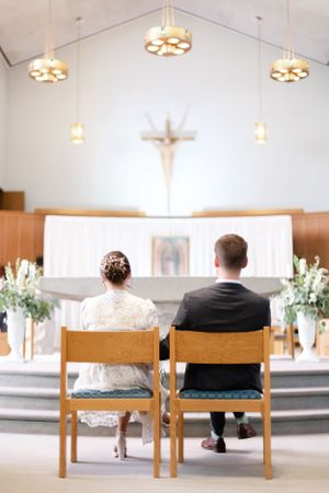 Church interior with wooden chairs and cross hanging on white wall with floral arrangements and chandelier lighting.