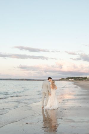 Dreamy beach wedding photo of couple walking along shore at sunset.