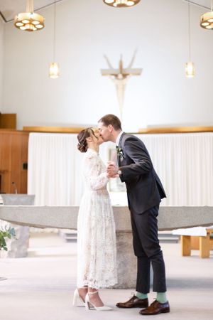 A couple shares a tender kiss during their wedding ceremony in a modern church with crosses and pendant lights.