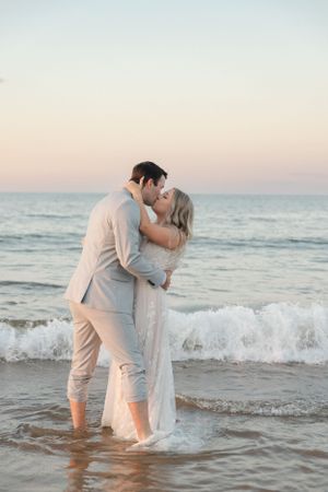 A romantic beach wedding scene with couple embracing at sunset in the gentle surf wearing light colored attire.