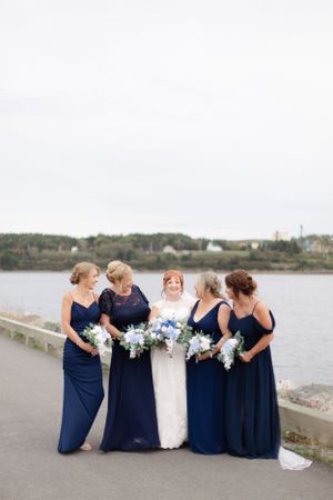 A bridal party in navy blue dresses poses along a waterfront path holding light blue and white floral bouquets.