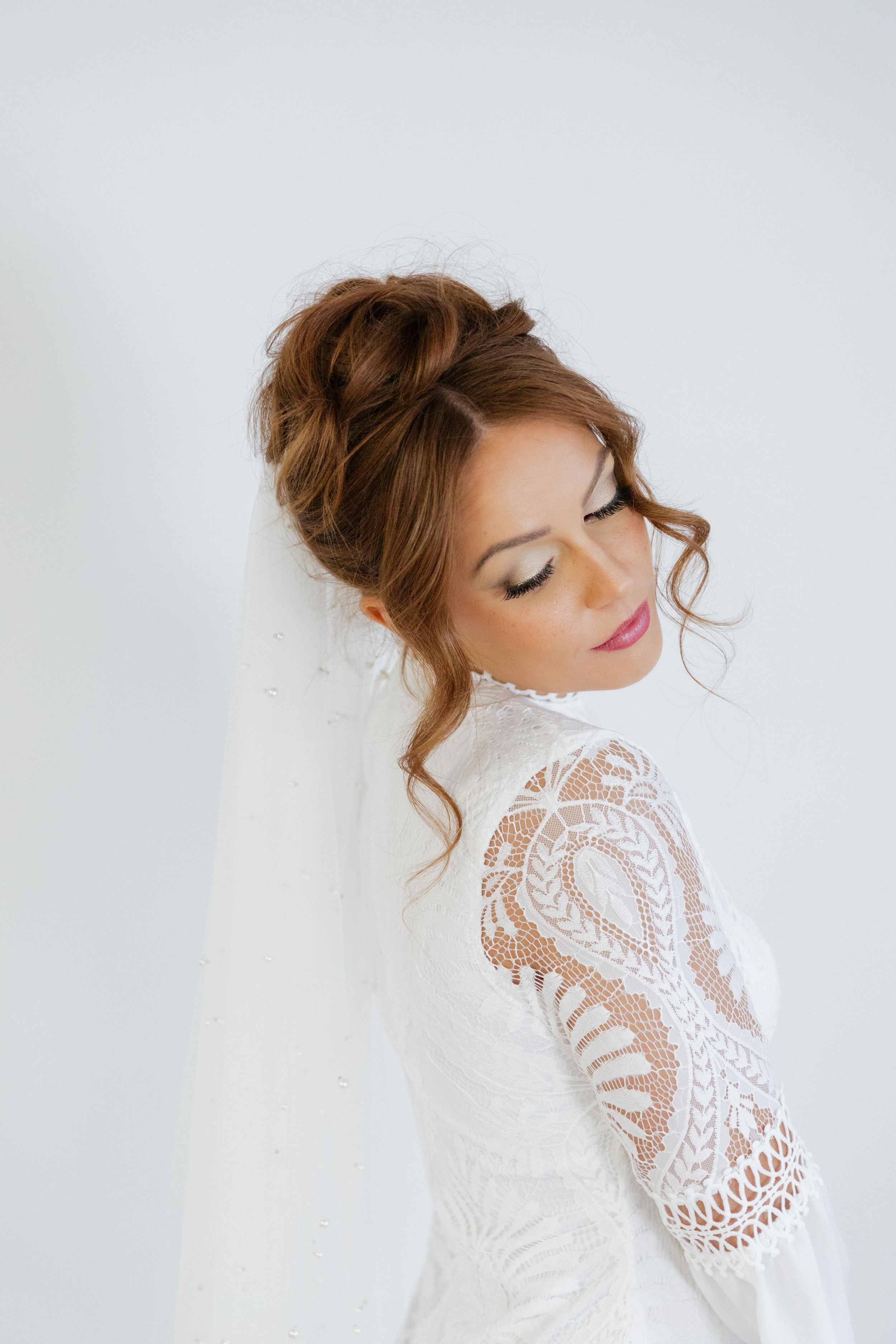 A bride in an elegant white lace long-sleeve wedding dress poses with eyes closed against a light background.