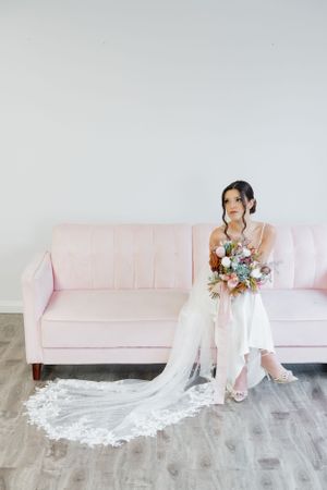 Elegant white wedding gown with lace train displayed by seated figure on blush pink sofa holding floral bouquet.