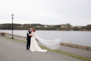 A wedding couple embraces on a waterfront path as the bride's veil flows gracefully in the breeze by a calm lake.