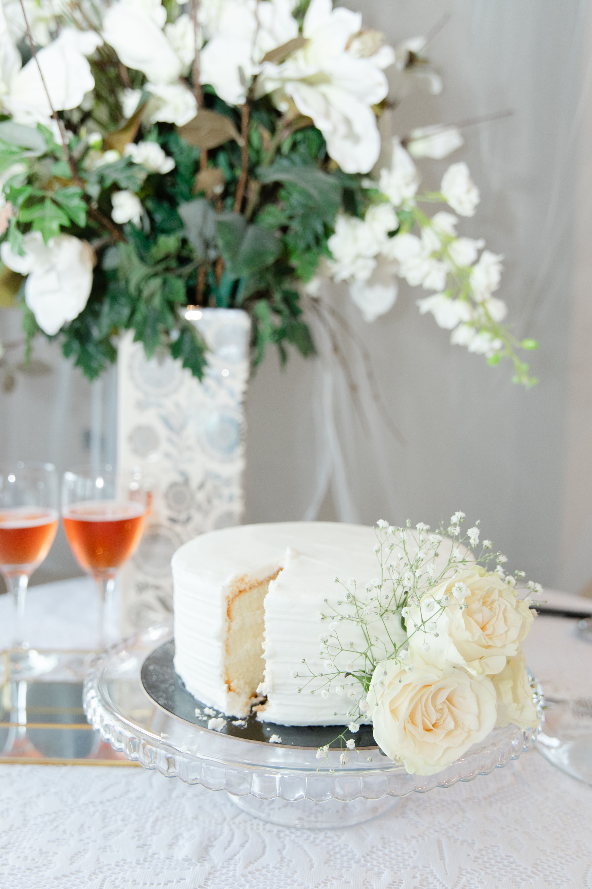 Elegant white wedding cake decorated with fresh roses and baby's breath on crystal cake stand with glasses of rosé.