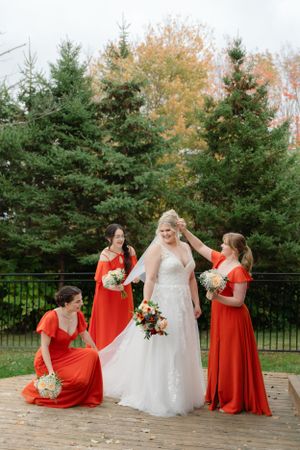 Bridal party poses outdoors with bridesmaids in vibrant coral dresses against evergreen trees.
