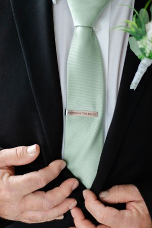Close up of hands adjusting a mint green necktie with a silver tie clip on a black suit jacket.