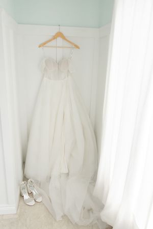 A white wedding gown hangs elegantly on a wooden hanger with matching bridal shoes placed below against a light wall.