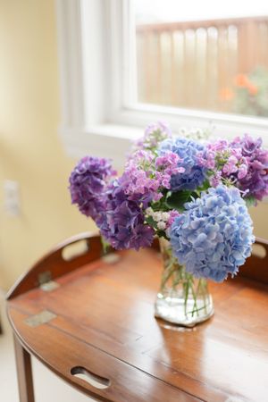 Beautiful arrangement of purple and blue hydrangeas in a glass vase on a wooden side table by a window.