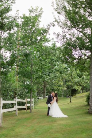 A couple shares a romantic moment on a grassy hillside with white fence and green trees framing the scenic outdoor setting.